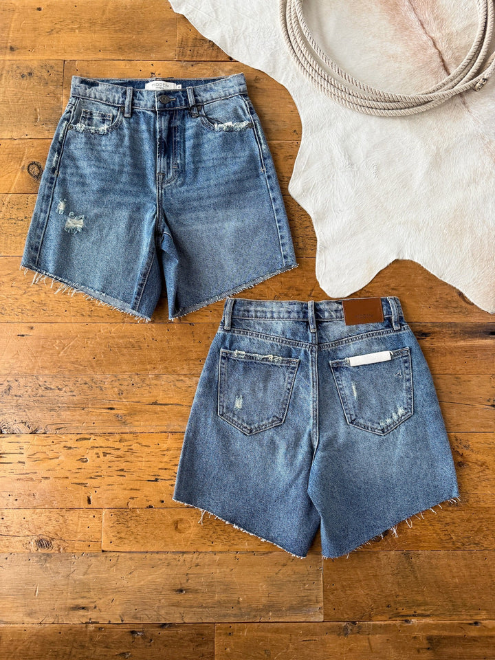 Two pairs of The Rowan Shorts by Savannah Sevens western life{&}style are laid flat on a wooden floor by a white cowhide rug and coiled rope, featuring raw hems and wear marks for a classic, well-loved look.