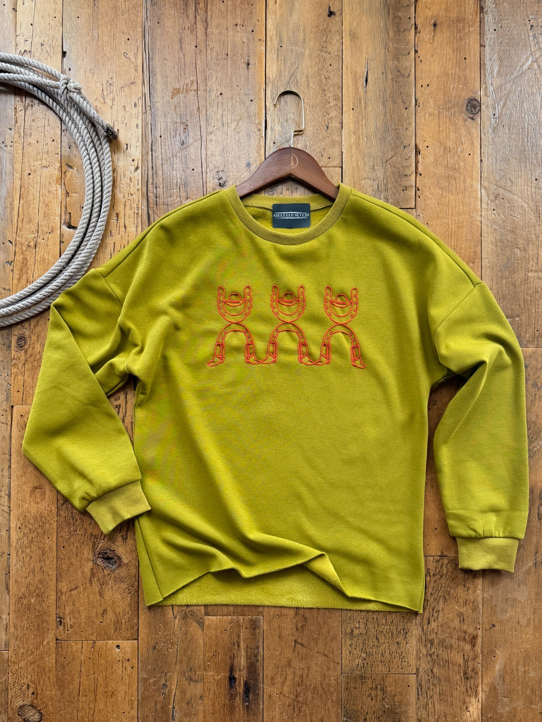Rowdy Chartreuse Embroidered Pullover in chartreuse with an orange embroidered design of three horseshoes on the chest, hanging on a wooden hanger against a wood-plank floor.