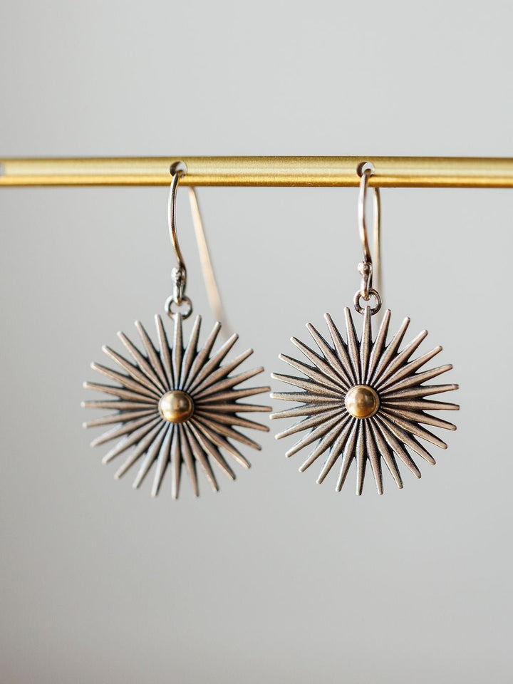 A pair of Rowel Earrings {Small} by Savannah Sevens western life{&}style, sterling silver with a sunburst design, radiating spikes, and a gold-colored center, hang from a gold bar against a neutral background.