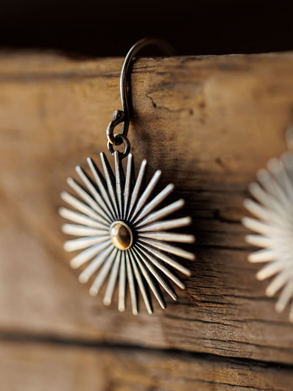 Close-up of Savannah Sevens western life{&}style Rowel Earrings {Small}—handmade sterling silver earrings with a sunburst shape featuring textured, radiating rays and hanging from a rustic wooden surface.