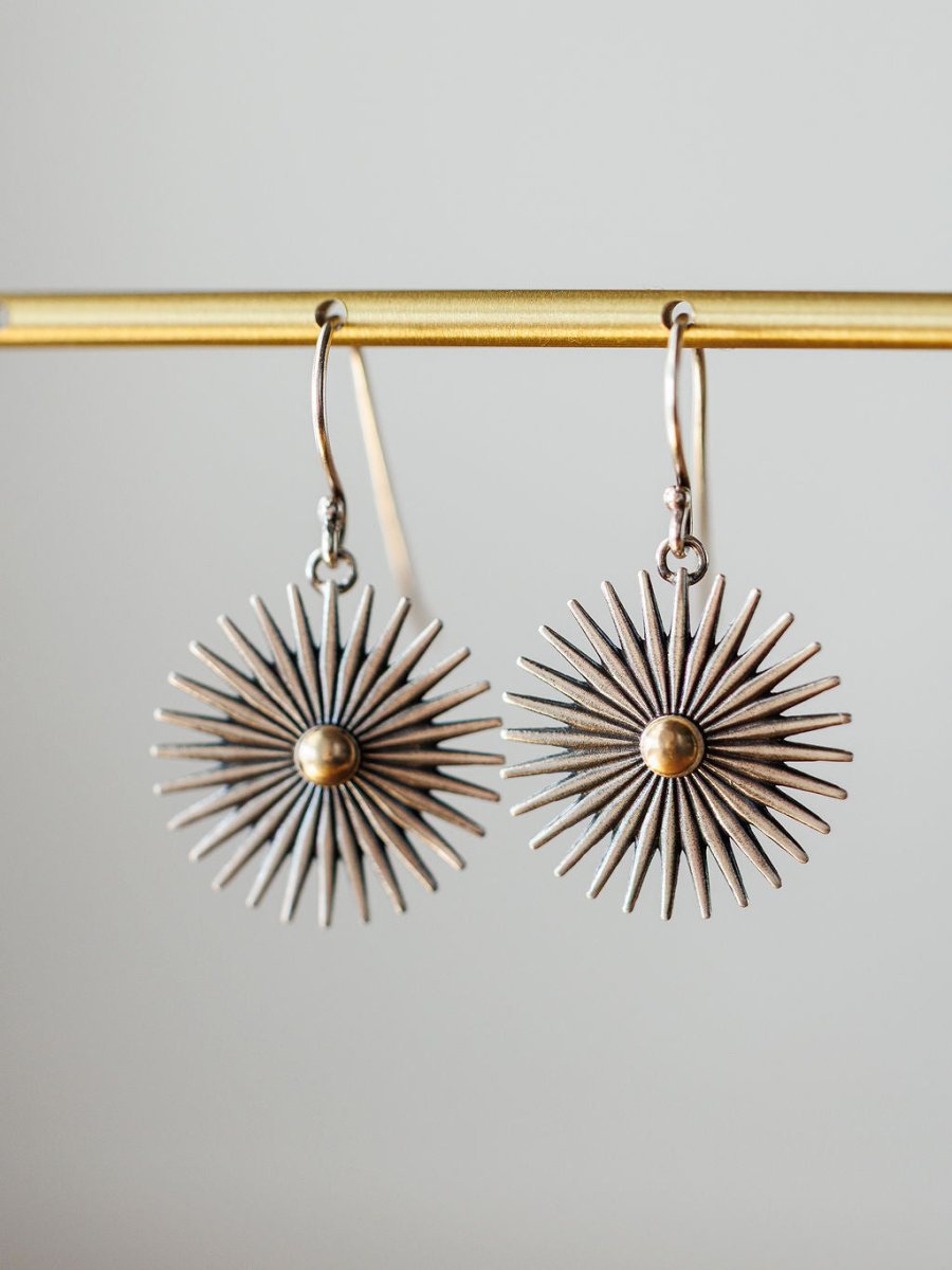 A pair of Rowel Earrings {Small} by Savannah Sevens western life{&}style, sterling silver with a sunburst design, radiating spikes, and a gold-colored center, hang from a gold bar against a neutral background.