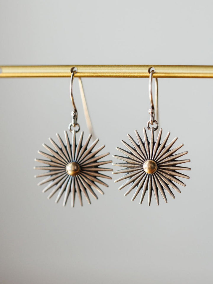 A pair of Rowel Earrings {Small} by Savannah Sevens western life{&}style, sterling silver with a sunburst design, radiating spikes, and a gold-colored center, hang from a gold bar against a neutral background.