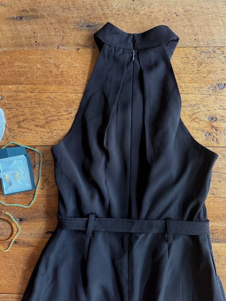 The Rowlett Black Sleeveless Jumpsuit by Savannah Sevens western life{&}style, featuring a halter neck and belt, is displayed on a wooden surface. Beside it are a gold beaded bracelet, a green beaded necklace, and a small blue box containing earrings.