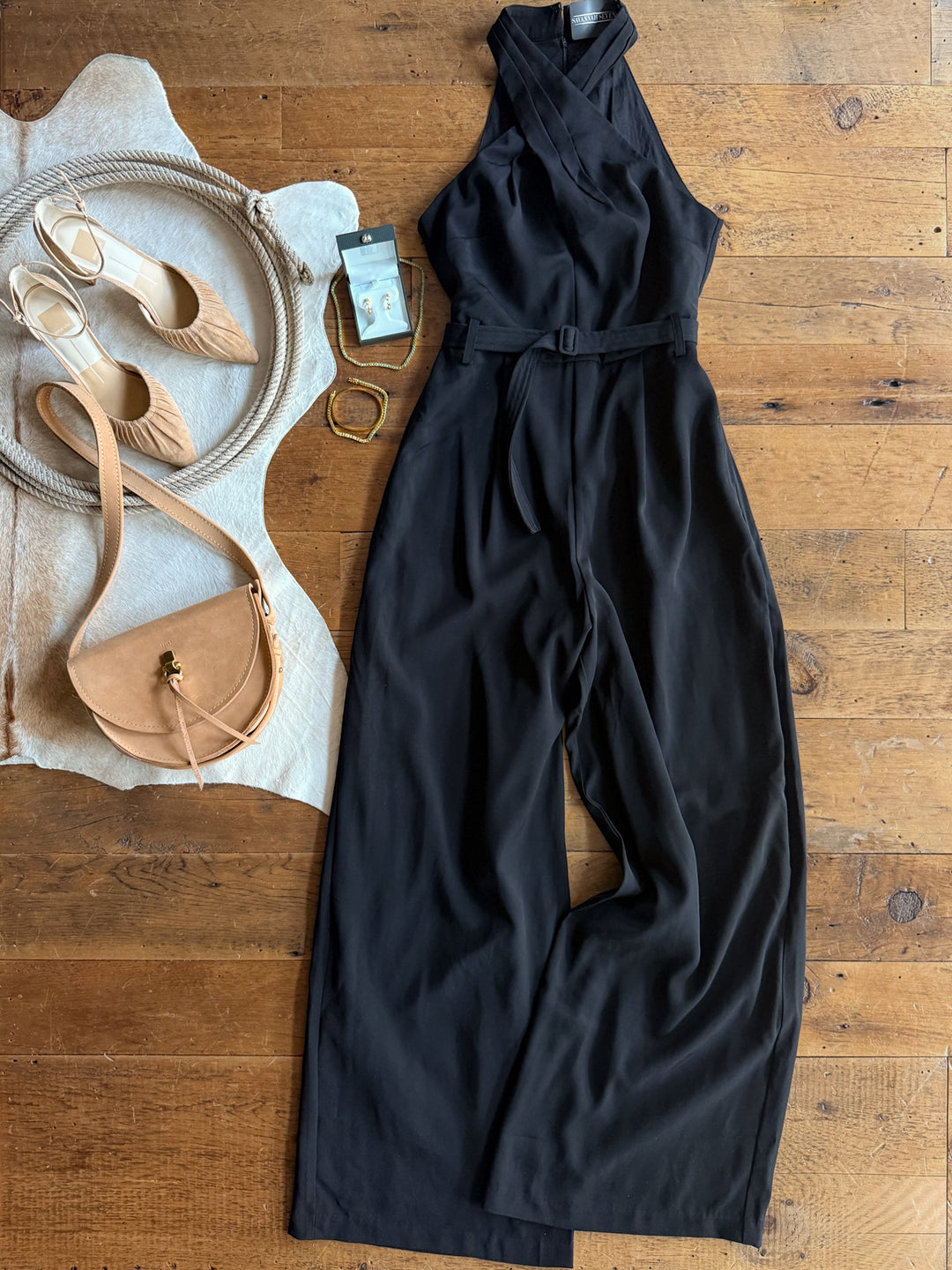 The Rowlett Black Sleeveless Jumpsuit from Savannah Sevens western life{&}style is styled on a wooden floor with beige heels, a tan crossbody bag, gold bracelets, a smartphone, and a round woven hat.