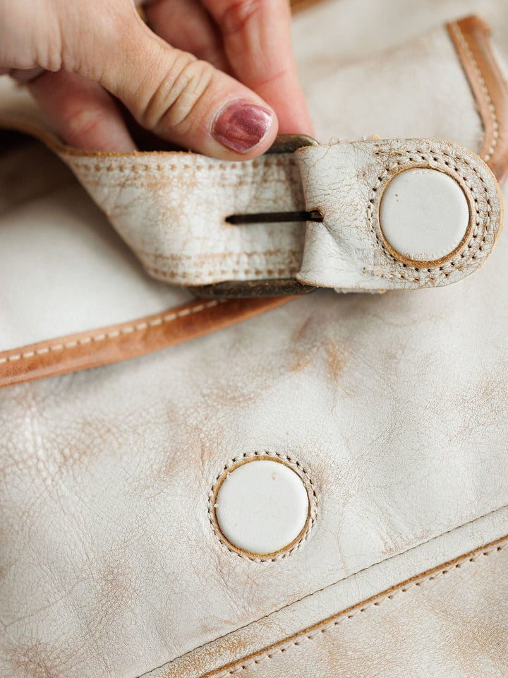 A hand with painted nails undoes the strap of a Savannah Sevens western life{&}style Rowley Leather Handbag, revealing its stitched button closure and distressed full grain leather texture.
