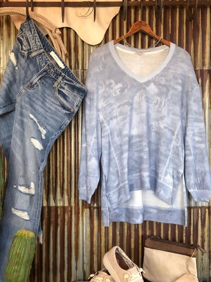 Savannah Sevens Roxton Watercolor Top in light blue, featuring a v-neck, long sleeves, an oversized fit, and a high-low hem, displayed next to distressed blue jeans.