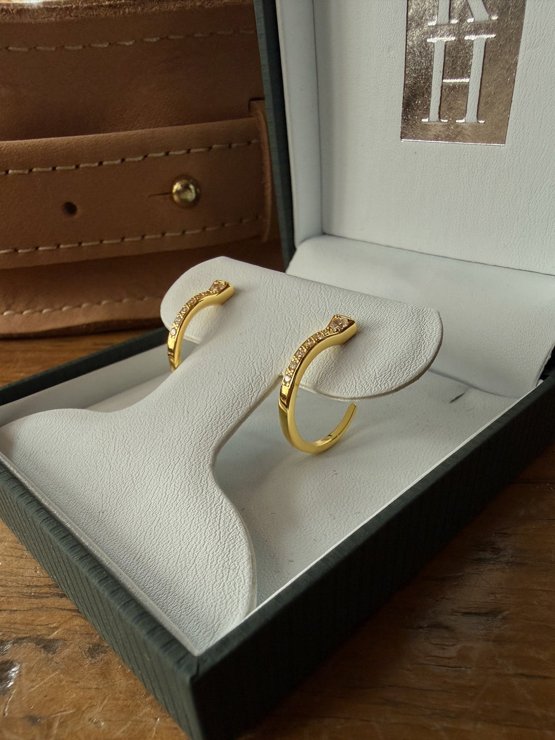 A pair of Rozel Gold Nail Hoop Earrings from Savannah Sevens western life{&}style is displayed in a white cushioned jewelry box, with a brown leather case partially visible in the background on a wooden surface.