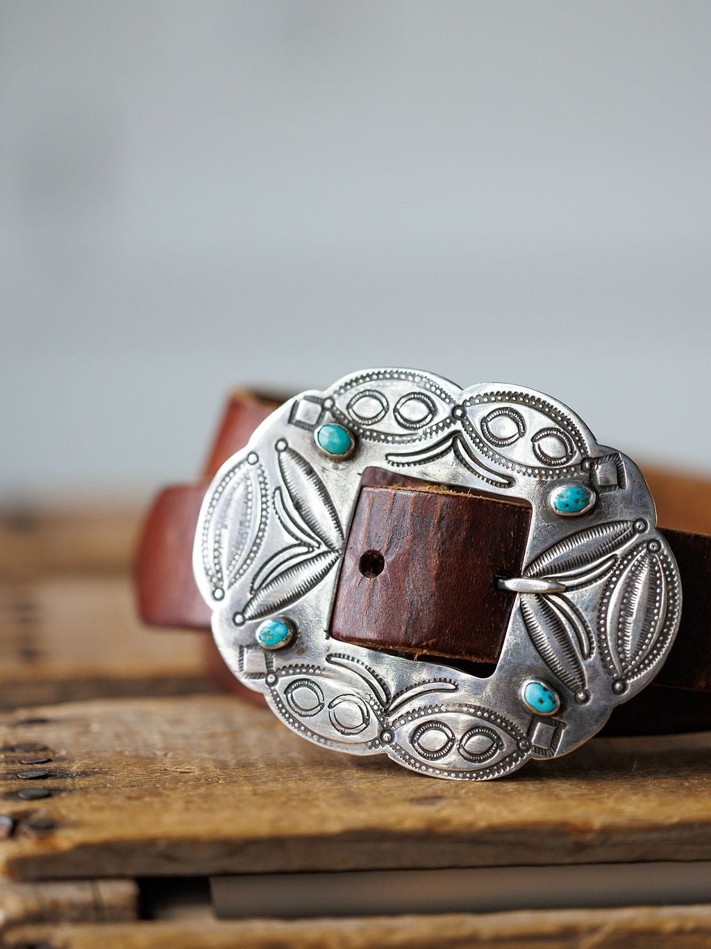 The Ruidoso Belt by Savannah Sevens western life{&}style is a brown leather belt with a Navajo-style engraved sterling silver buckle and four turquoise stones, displayed on a wooden surface.