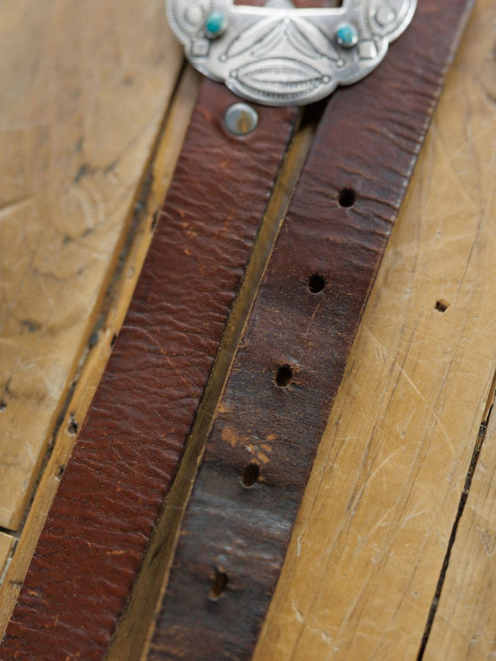 Close-up of the Ruidoso Belt by Savannah Sevens western life{&}style, a worn brown leather belt with multiple holes resting on wood. A partially visible Navajo sterling silver and turquoise buckle is seen at the top.
