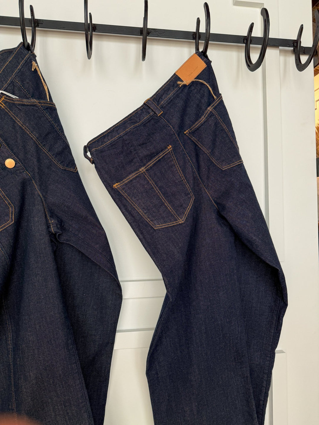 Two pairs of dark wash denim jeans with tan stitching hang on black hooks against a white paneled door. Fully shown on the right is The Rylan High Rise Wide Leg Jean by Savannah Sevens western life{&}style.