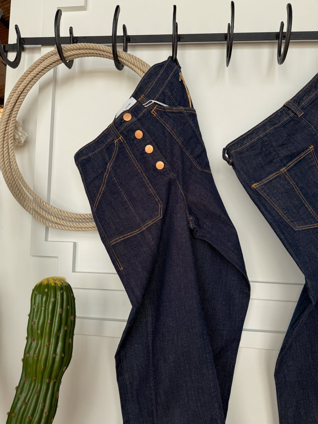 The Rylan High Rise Wide Leg Jean by Savannah Sevens western life{&}style, featuring a dark wash and four copper buttons, hangs on a black hook rail beside a green cactus and coiled rope against a white paneled wall.