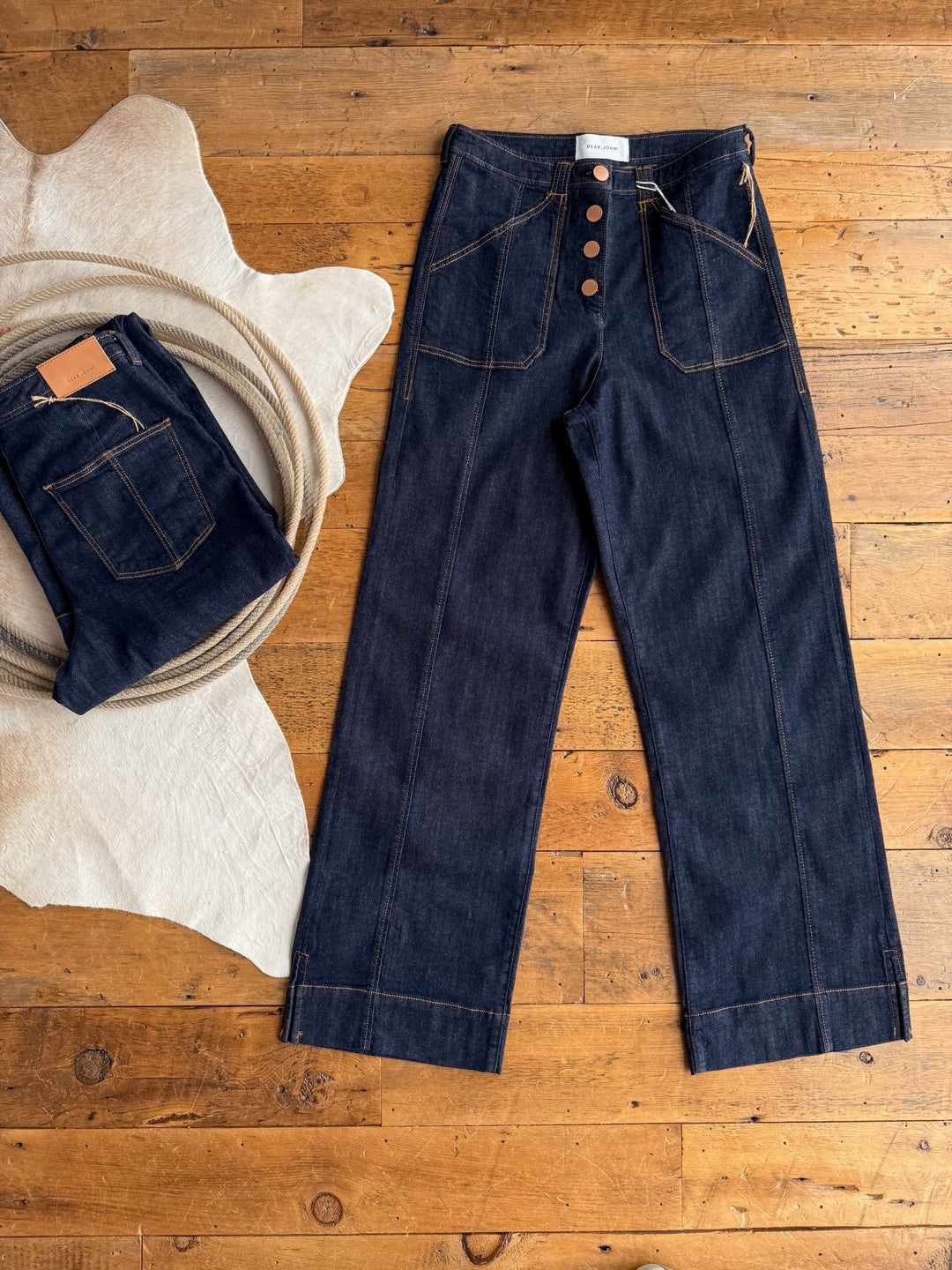 A pair of Savannah Sevens western life{&}style's The Rylan High Rise Wide Leg Jean in dark wash with tan stitching and large front pockets lies flat on a wooden floor, beside a folded pair on a cream-colored cloth.