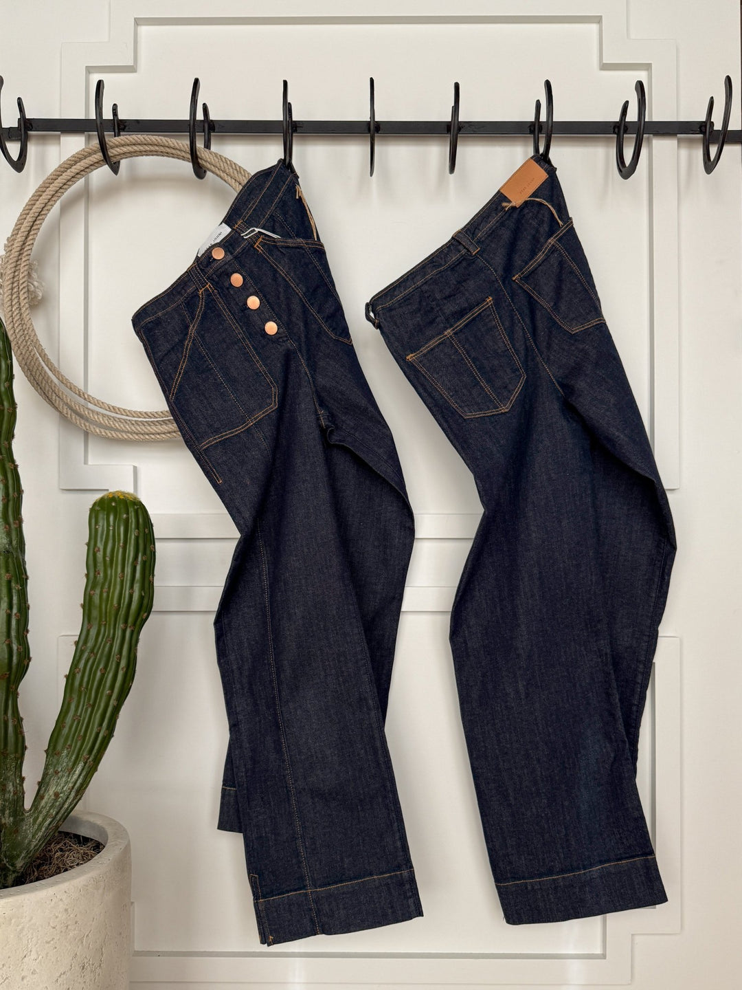 Two pairs of The Rylan High Rise Wide Leg Jeans by Savannah Sevens western life{&}style hang side by side on black hooks against a white door, with a potted cactus to the left and a decorative rope ring hanging on the door.