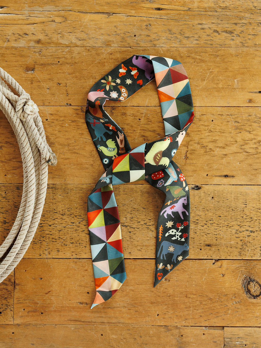 A Farm Folk Silk Twilly by Savannah Sevens western life&style, a long, narrow silk western tie scarf with a geometric print on one side and a whimsical folk-art animal pattern on the other, tied in a knot on a wooden surface next to a rope.