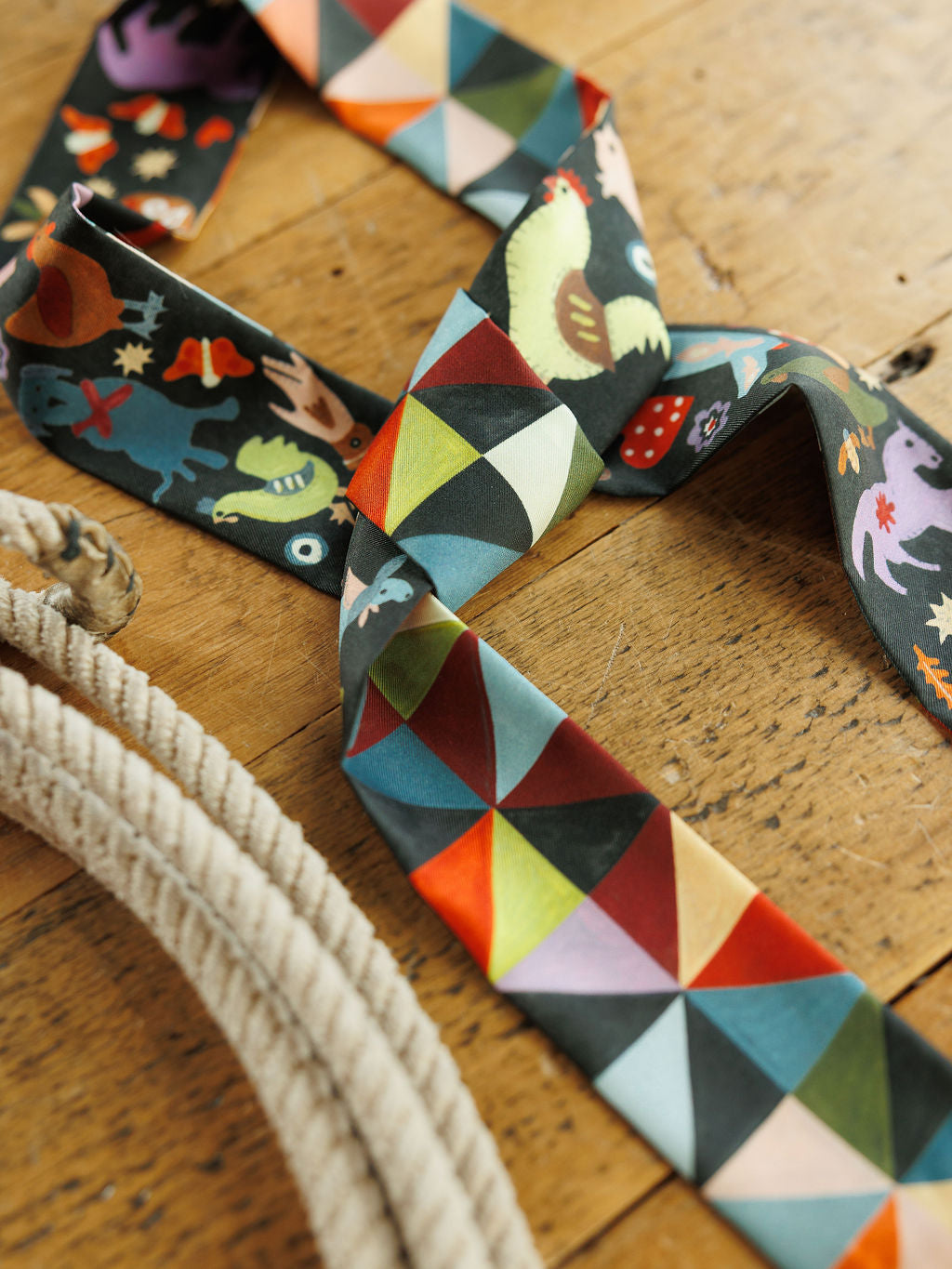 A Farm Folk Silk Twilly by Savannah Sevens western life&style, featuring a long, thin silk tie scarf with colorful geometric triangles on one side and a whimsical folk art animal print on the other, resting on a wooden surface beside a coiled rope.