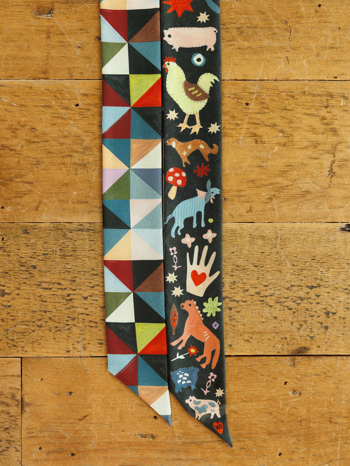 Two long, thin Farm Folk Silk Twilly scarves lie side-by-side: one with a multicolored geometric print and the other featuring a dark background with illustrated animals, a heart-adorned hand, and mushrooms.