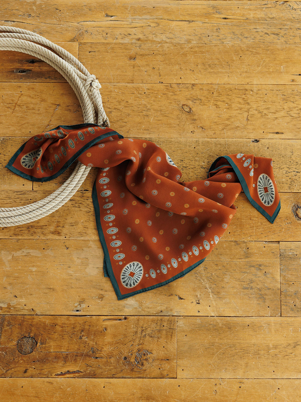 San Miguel Silk Scarf by Savannah Sevens western life&style in rust-colored silk with yellow circles, geometric motifs, and a dark teal border, resting on a wooden surface next to a coiled rope.