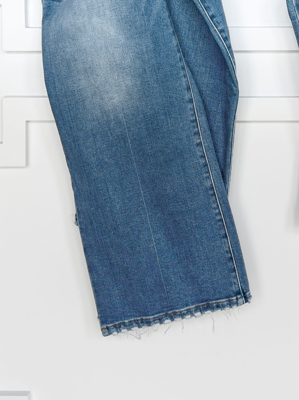 A close-up of the lower leg of Savannah Sevens western life{&}style’s The Paxton High Rise Wide Leg Jean in blue stretch denim, featuring a frayed hem, hanging against a white background.