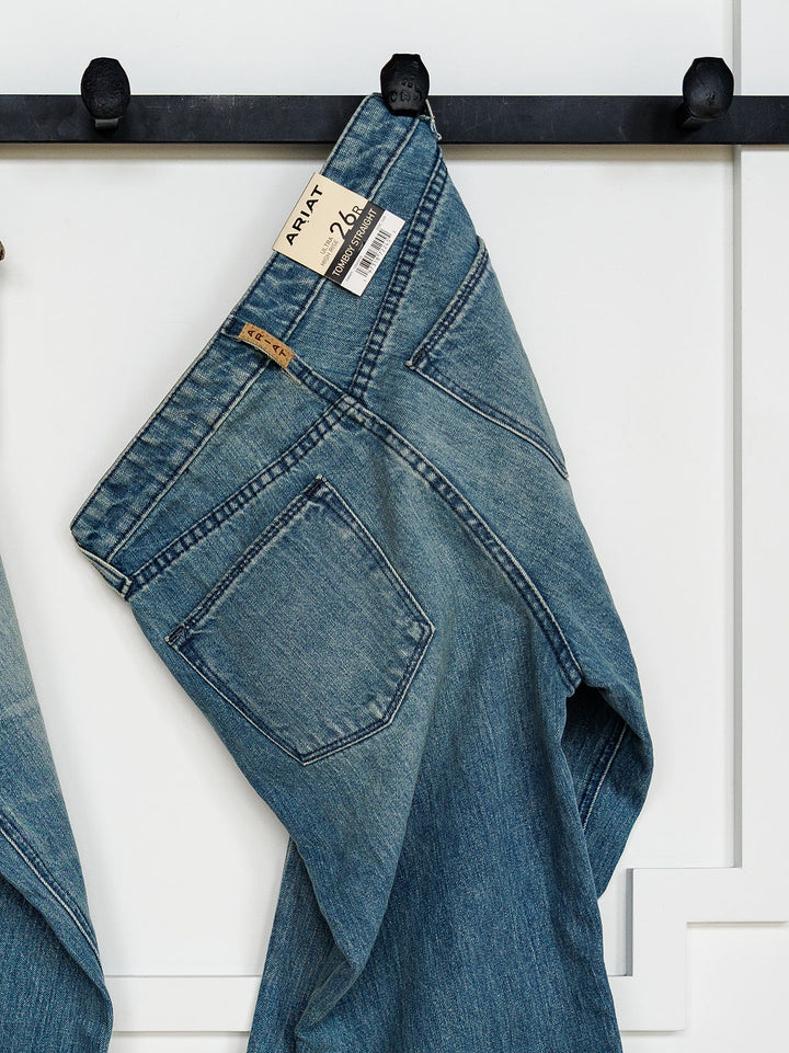 A pair of Ariat The Truett High Rise Relaxed Straight Leg Jeans with a visible brand tag hangs by the waistband on a black wall hook against a white paneled background.