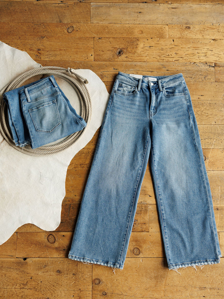The Paxton High Rise Wide Leg Jean by Savannah Sevens western life{&}style is laid flat on a wooden floor, beside a coiled rope and another folded pair of jeans on a white cowhide rug.