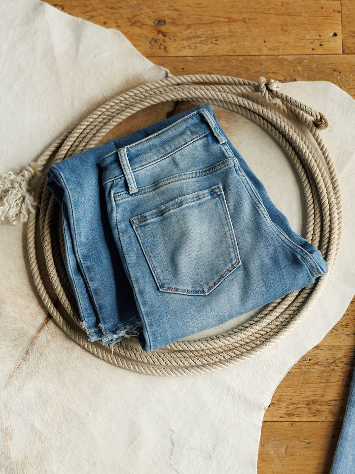 The Paxton High Rise Wide Leg Jean by Savannah Sevens western life{&}style in light blue, folded neatly on a coiled rope over a pale animal hide, displayed atop a wooden floor.