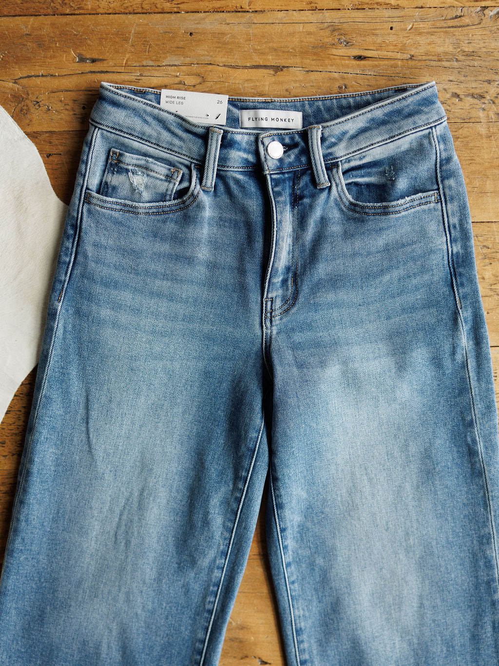 The Paxton High Rise Wide Leg Jean by Savannah Sevens western life{&}style in light blue stretch denim with slight fading and button closure, laid flat on wood. A white fabric or paper appears partially visible on the left.