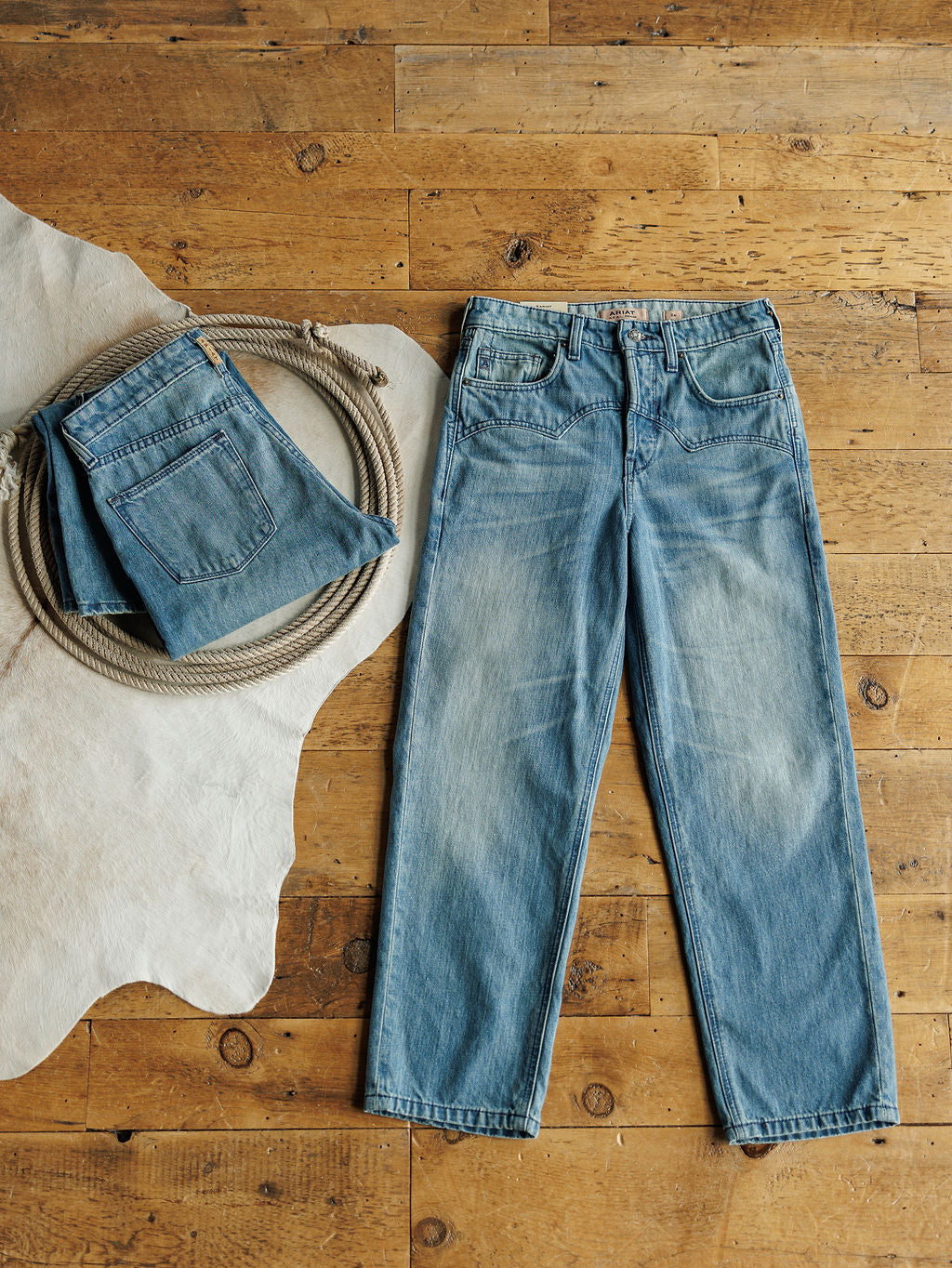 A pair of Ariat Truett High Rise Relaxed Straight Leg Jeans in denim blue is laid flat on a wooden floor beside a coiled rope and another folded pair on a white animal hide rug.