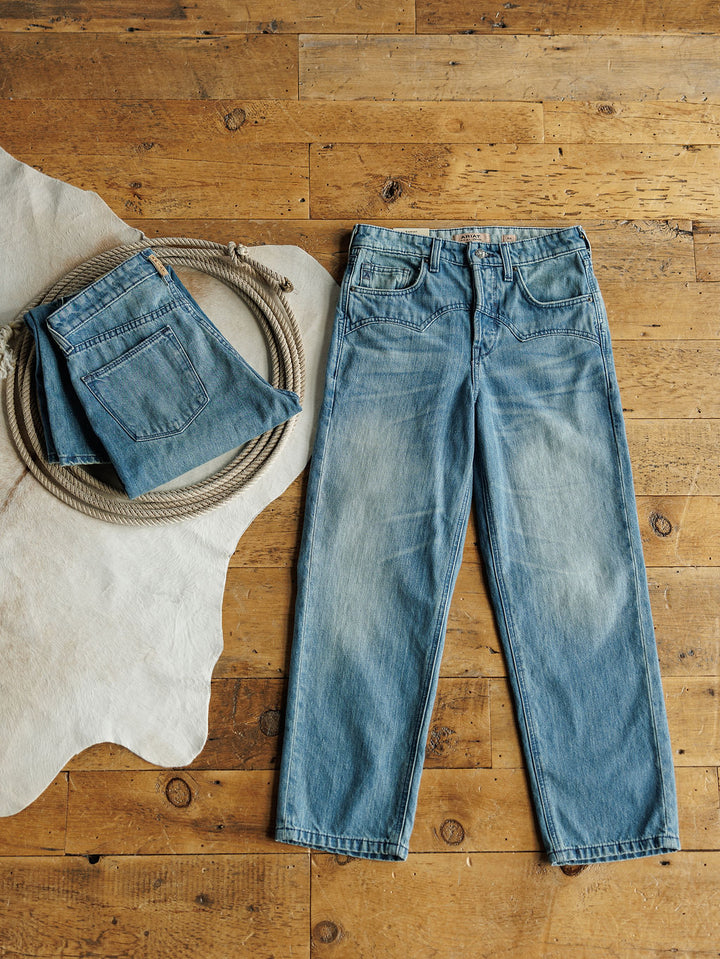 A pair of Ariat Truett High Rise Relaxed Straight Leg Jeans in denim blue is laid flat on a wooden floor beside a coiled rope and another folded pair on a white animal hide rug.