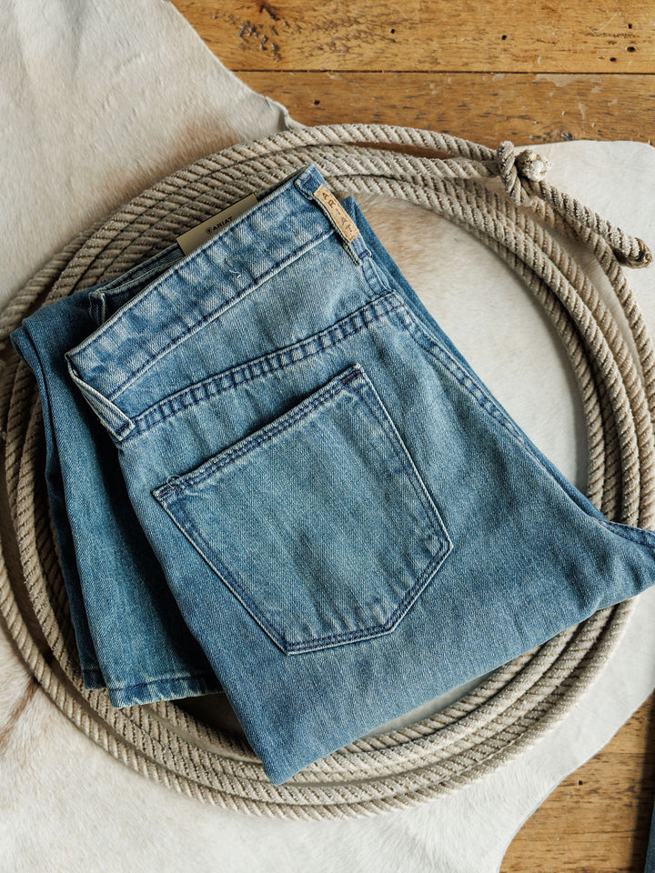 A folded pair of Ariat's The Truett High Rise Relaxed Straight Leg Jeans sits on a coiled rope atop a white cowhide rug, set against a wooden floor backdrop.