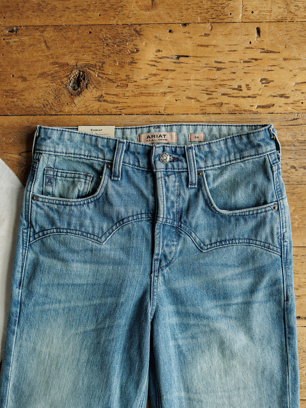 The Ariat Truett High Rise Relaxed Straight Leg Jean in medium wash, featuring front pockets, visible stitching, a button closure, and the Ariat label on the waistband, laid flat on a rustic wooden surface.