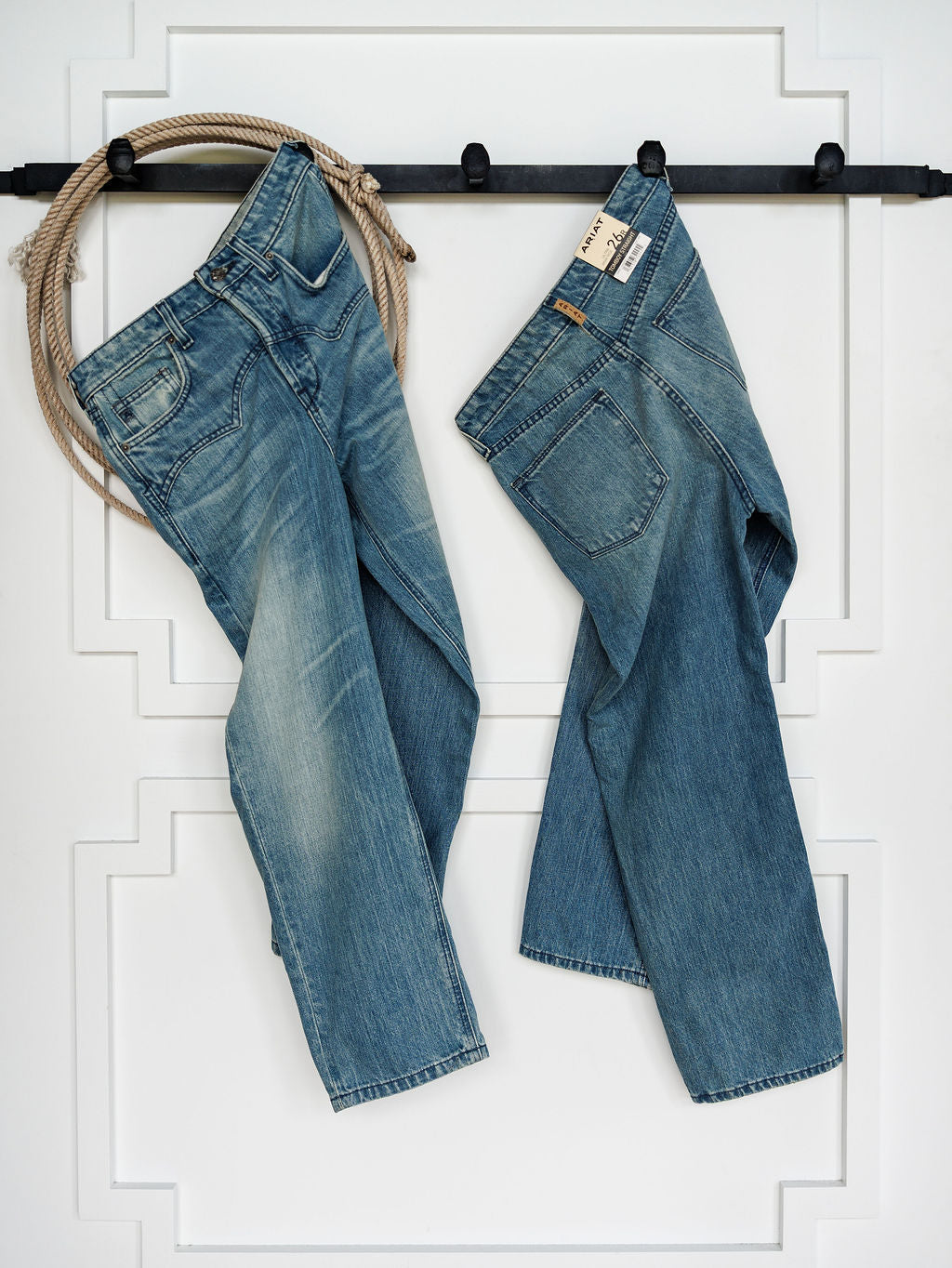Two pairs of medium wash blue jeans, including Ariat's The Truett High Rise Relaxed Straight Leg Jean, hang from a black wall-mounted rack against white paneling. A coiled rope sits at the left end, and one pair displays Ariat denim tags.
