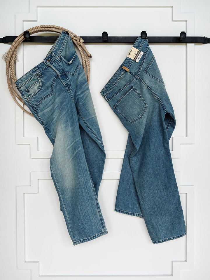 Two pairs of medium wash blue jeans, including Ariat's The Truett High Rise Relaxed Straight Leg Jean, hang from a black wall-mounted rack against white paneling. A coiled rope sits at the left end, and one pair displays Ariat denim tags.
