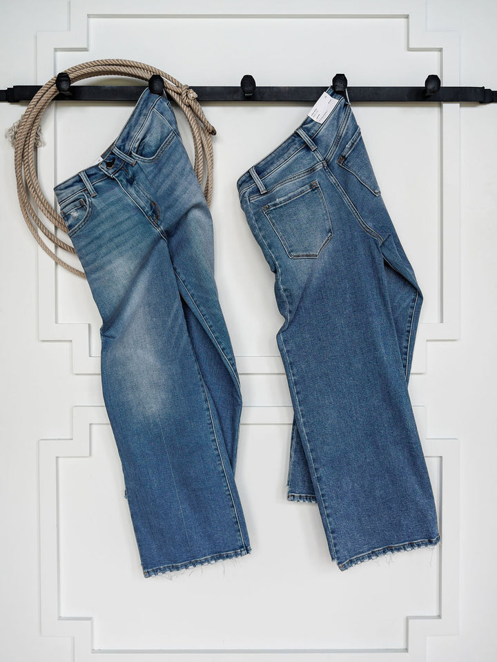 Two pairs of The Paxton High Rise Wide Leg Jeans by Savannah Sevens western life{&}style with a faded wash and frayed hems hang from a black rod against a white wall, with a coil of rope looped over the left side.