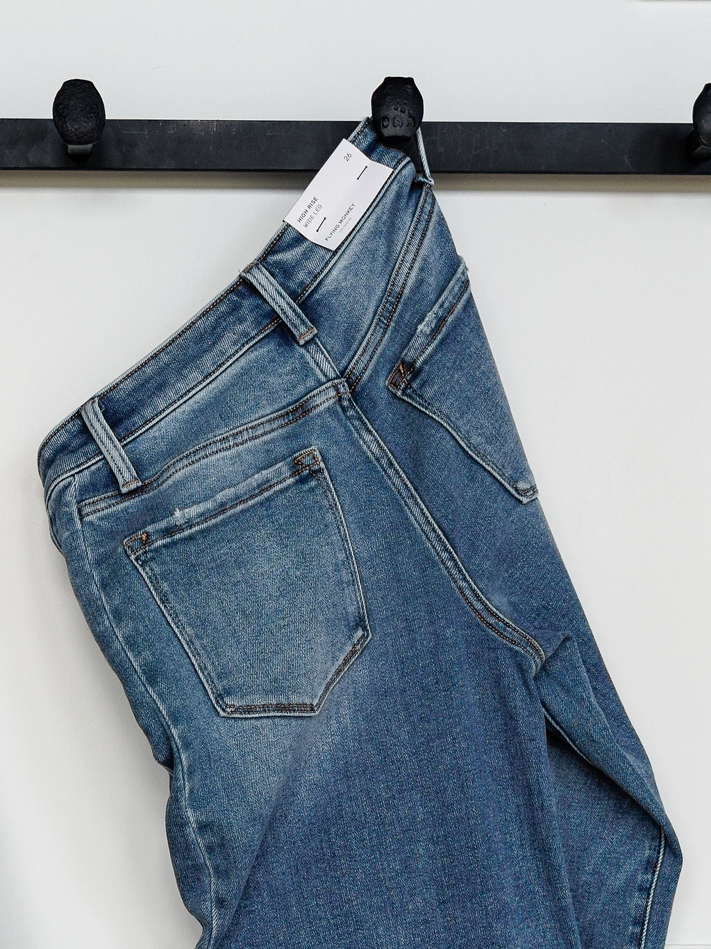The Paxton High Rise Wide Leg Jean by Savannah Sevens western life{&}style, in blue stretch denim with a white tag, hangs on a black metal hook against a plain white wall.