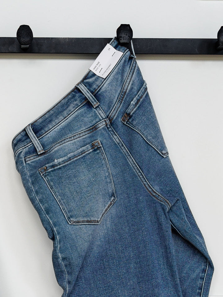 The Paxton High Rise Wide Leg Jean by Savannah Sevens western life{&}style, in blue stretch denim with a white tag, hangs on a black metal hook against a plain white wall.