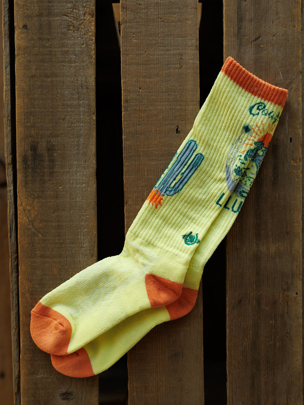 The Desert Cowgirl Crew Socks by Savannah Sevens western life{&}style feature a yellow base with orange toes and heels, blue cactus, and desert scene, displayed on rustic wood.