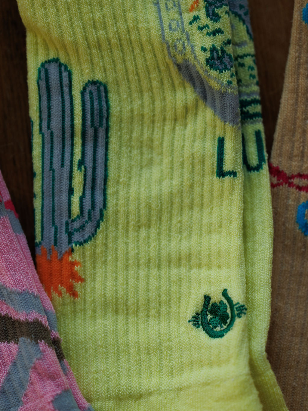 A close-up of Savannah Sevens western life{&}style’s Desert Cowgirl Crew Socks in yellow, featuring embroidered cactus and horseshoe designs, with green accents and glimpses of other colorful socks at the edges.