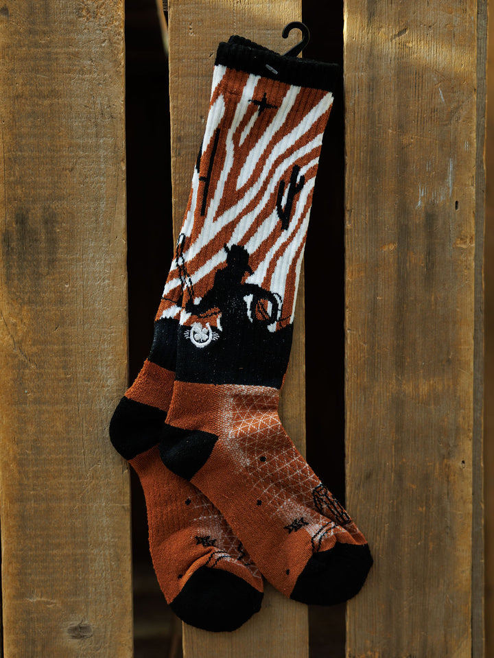 A pair of Maverick Crew Socks by Savannah Sevens western life{&}style, featuring orange, black, and white zebra stripes plus cactus and bicycle rider designs, hang on a wooden fence.