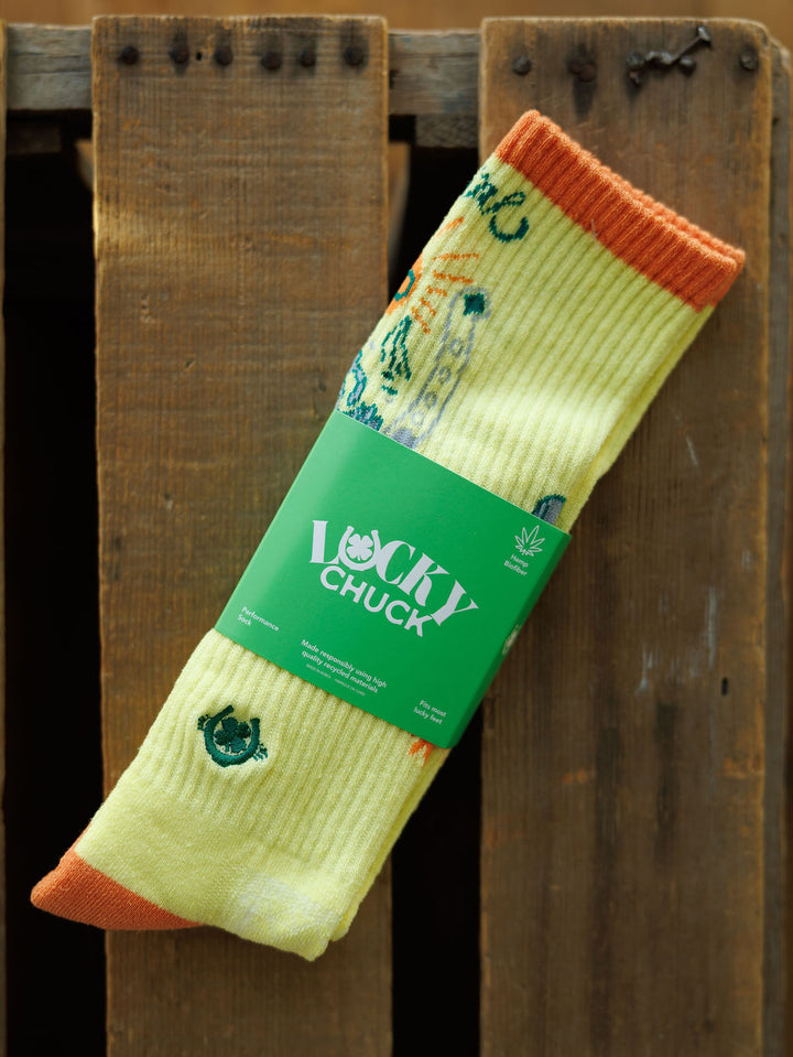 A pair of Desert Cowgirl Crew Socks by Savannah Sevens western life{&}style, yellow with orange toes and cuffs plus colorful embroidery, is wrapped in a green Lucky Chuck label and set on a wooden surface.