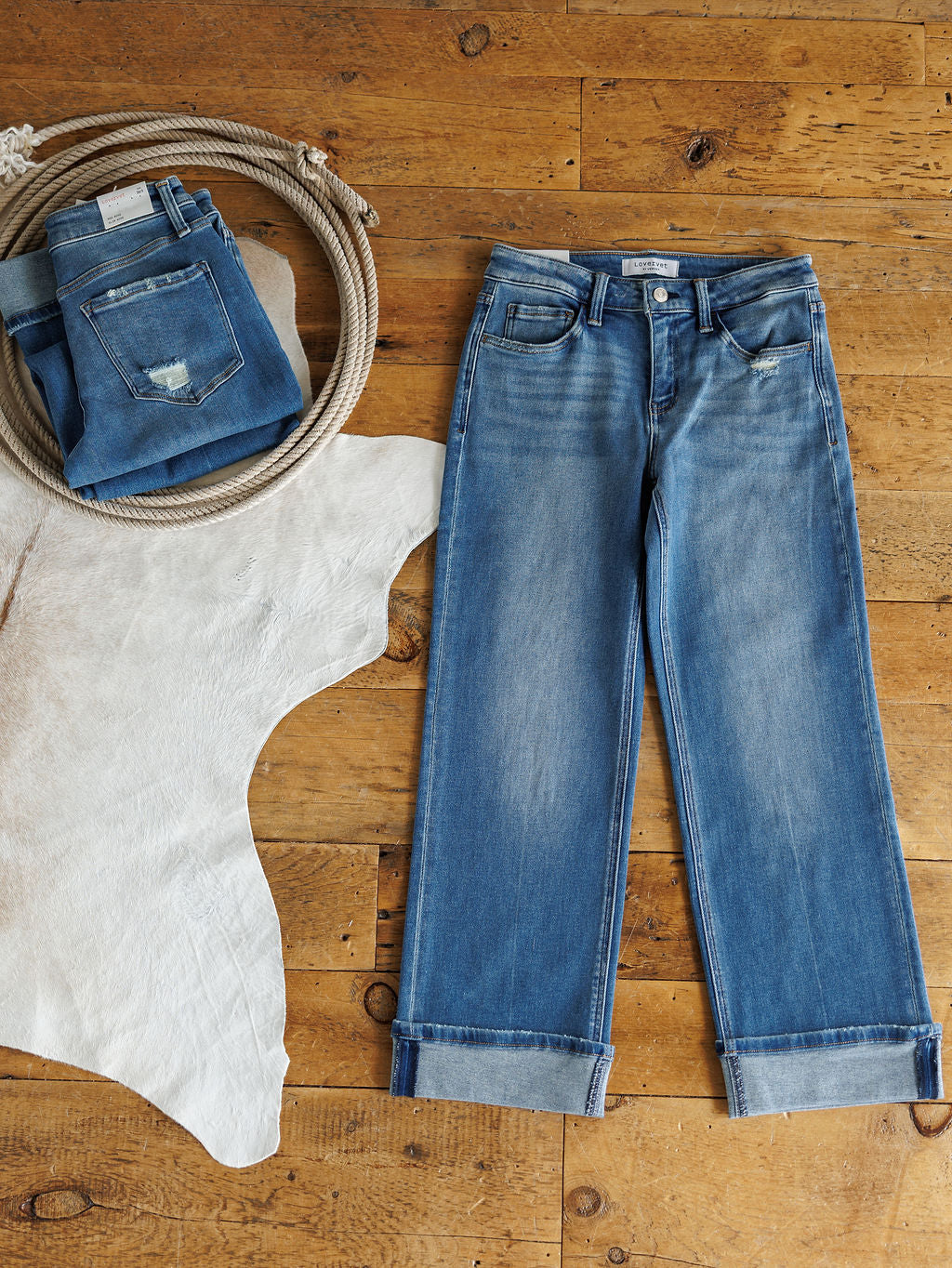 The Mack High Rise Relaxed Straight Leg Jean by Savannah Sevens western life{&}style is laid flat on wood next to folded Vervet jeans on a white cowhide rug and a coiled rope, adding a rustic, western touch.