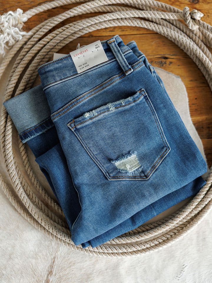 The Mack High Rise Relaxed Straight Leg Jean by Savannah Sevens western life{&}style, a medium wash pair with a frayed back pocket, is neatly folded on a wooden surface beside coiled rope and white fabric.