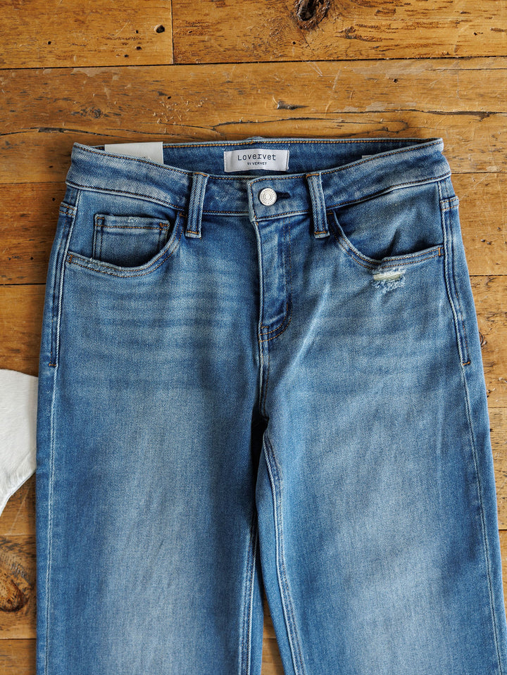 The Mack High Rise Relaxed Straight Leg Jean by Savannah Sevens western life{&}style features blue denim with slight fading and a small distressed patch near the right pocket, displayed flat on rustic wood with a partially visible white fabric on the left.