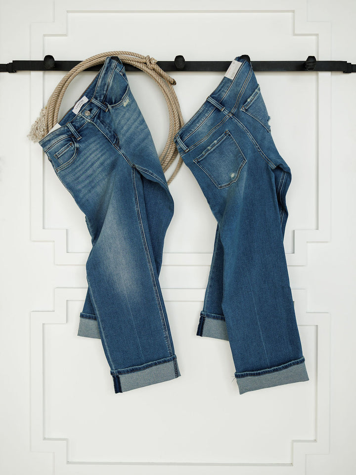 Two pairs of The Mack High Rise Relaxed Straight Leg Jean by Savannah Sevens western life{&}style in medium wash, with rolled cuffs, hang from a black rod against a white wall. A coiled rope is draped over the rod beside the jeans.