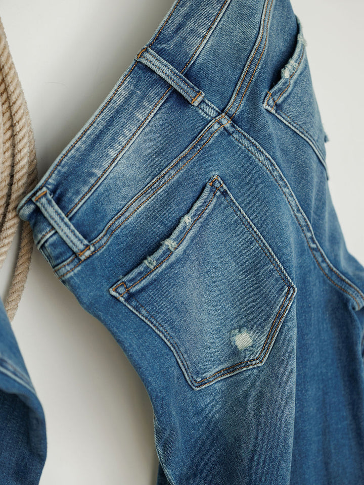 Close-up of The Mack High Rise Relaxed Straight Leg Jean by Savannah Sevens western life{&}style in medium wash, showing worn edges and a small distressed patch on the back pocket. Thick rope visible on the left.