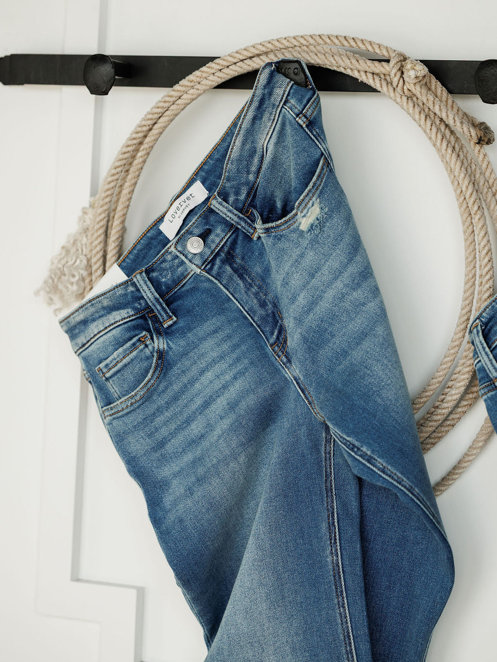 A pair of Savannah Sevens western life{&}style The Mack High Rise Relaxed Straight Leg Jeans in medium wash hangs on a black hook, with a coiled rope behind them against a white wall.