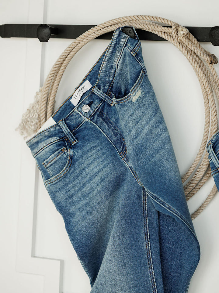 A pair of Savannah Sevens western life{&}style The Mack High Rise Relaxed Straight Leg Jeans in medium wash hangs on a black hook, with a coiled rope behind them against a white wall.