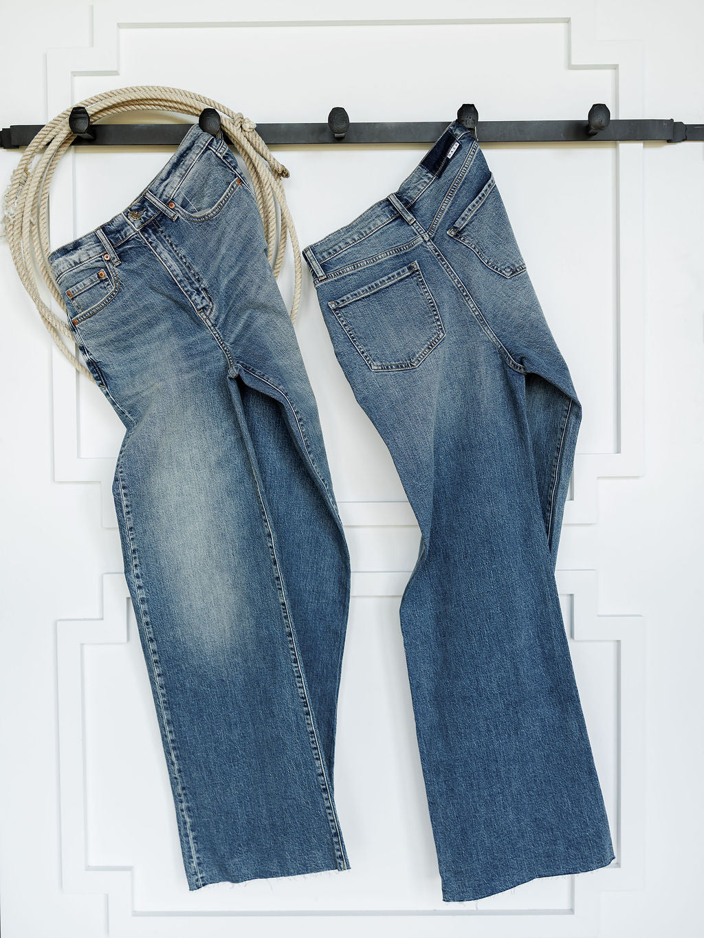 Two pairs of The Leyden High Rise Wide Leg Jean by Savannah Sevens western life{&}style hang on a black wall hook, with a coiled rope draped over the left hook. A white wall with decorative molding serves as the background.