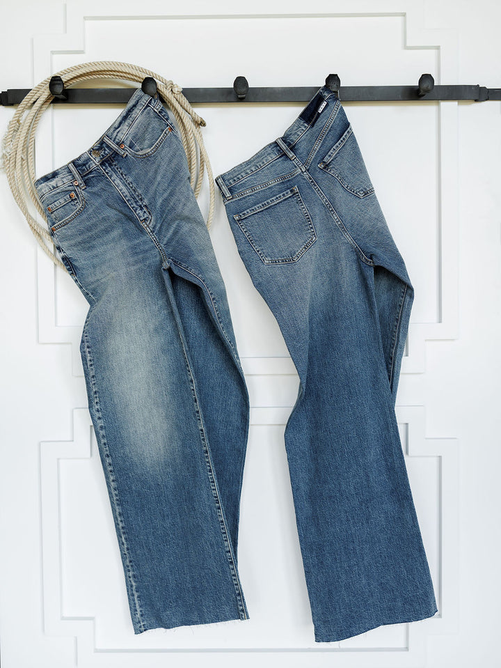 Two pairs of The Leyden High Rise Wide Leg Jean by Savannah Sevens western life{&}style hang on a black wall hook, with a coiled rope draped over the left hook. A white wall with decorative molding serves as the background.
