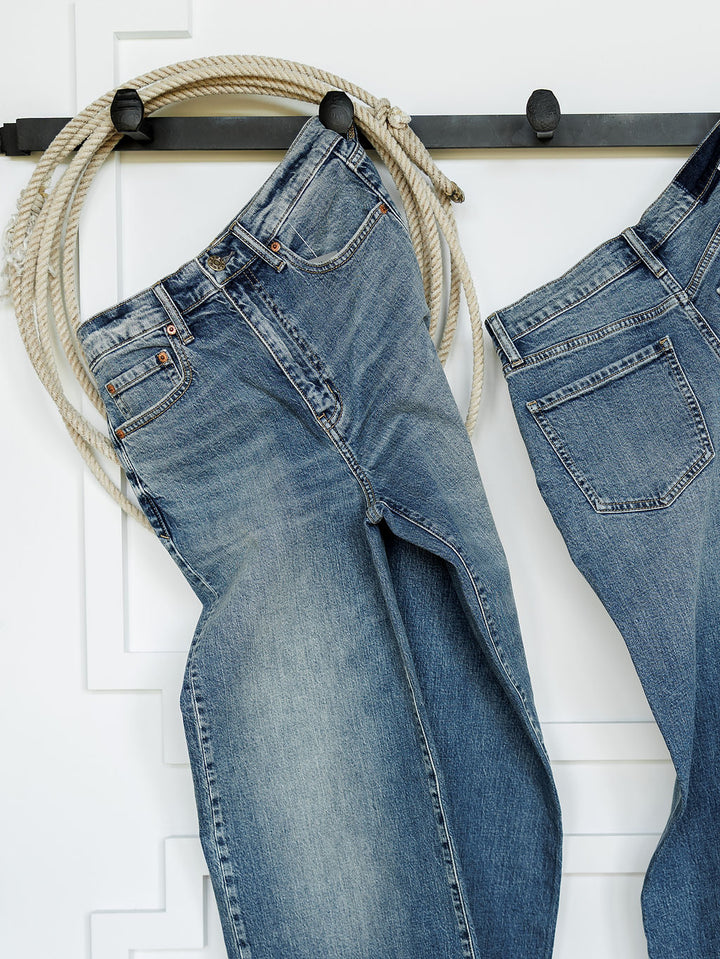 Two pairs of The Leyden High Rise Wide Leg Jeans by Savannah Sevens western life{&}style in medium wash denim hang side by side from black hooks on a white wall, with a coiled rope draped above them.
