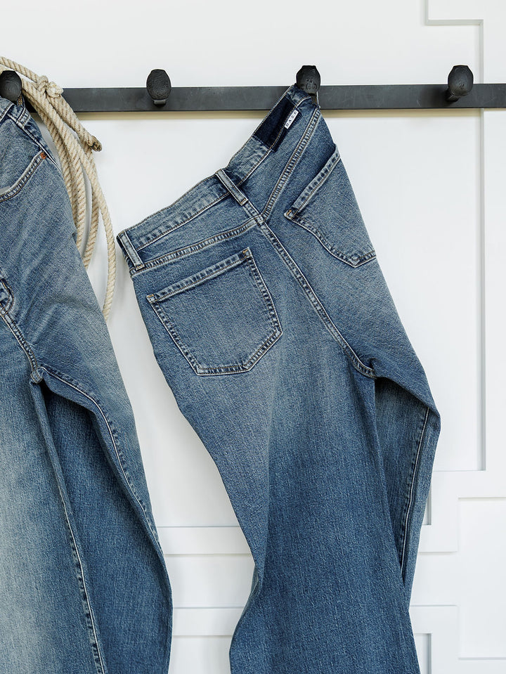 The Leyden High Rise Wide Leg Jean by Savannah Sevens western life{&}style in medium wash denim hangs on black hooks against a white wall, alongside another pair of jeans and a coiled rope.